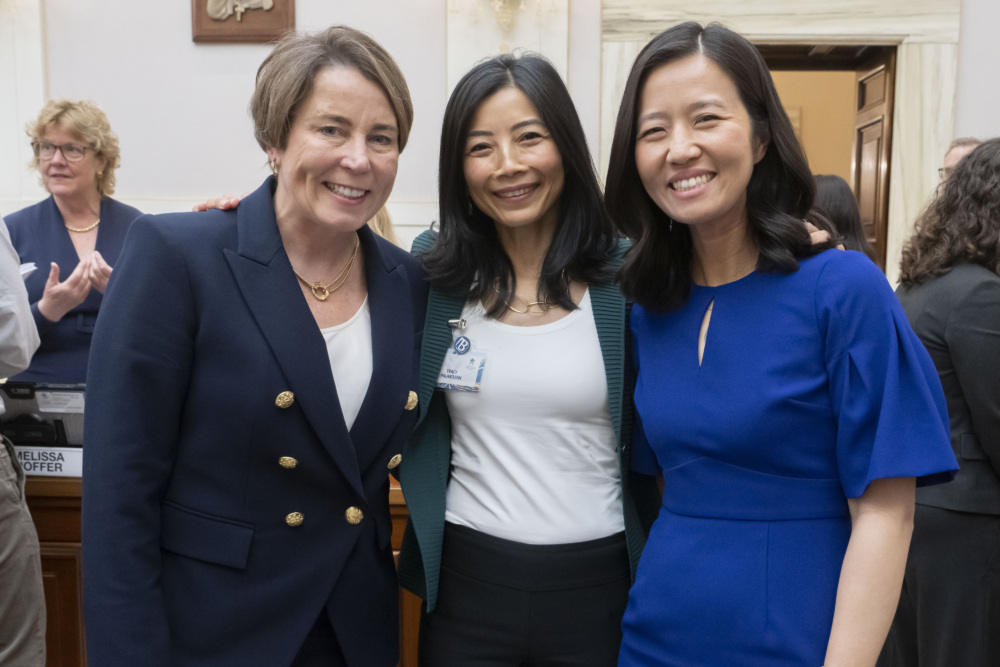 Three women pose for a photo in a formal indoor setting, each smiling at the camera. The woman on the left is wearing a navy blazer, the middle woman wears a green cardigan, and the woman on the right wears a blue dress.